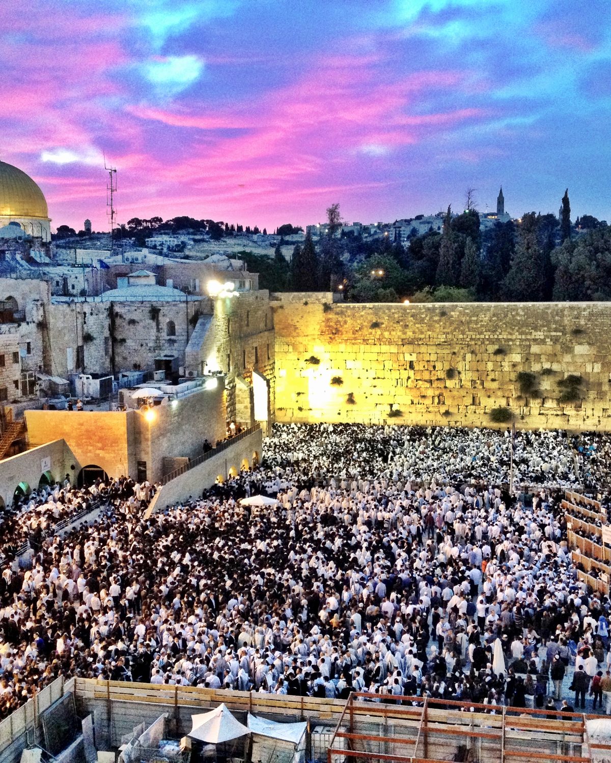 The Western Wall in Jerusalem under a pink sunset sky, with the golden Dome of the Rock above